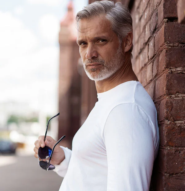 Mature man leaning against a brick wall, holding sunglasses and wearing a white long-sleeved shirt, looking confidently at the camera - Facial Fat Grafting in Portsmouth, NH