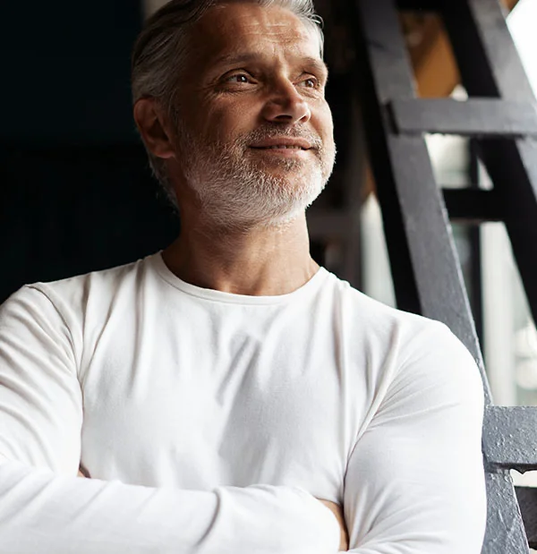 A mature man with silver hair and a beard, wearing a white long-sleeve shirt. He is smiling, looking off to the side, with his arms crossed - Lip Lift in Portsmouth, NH