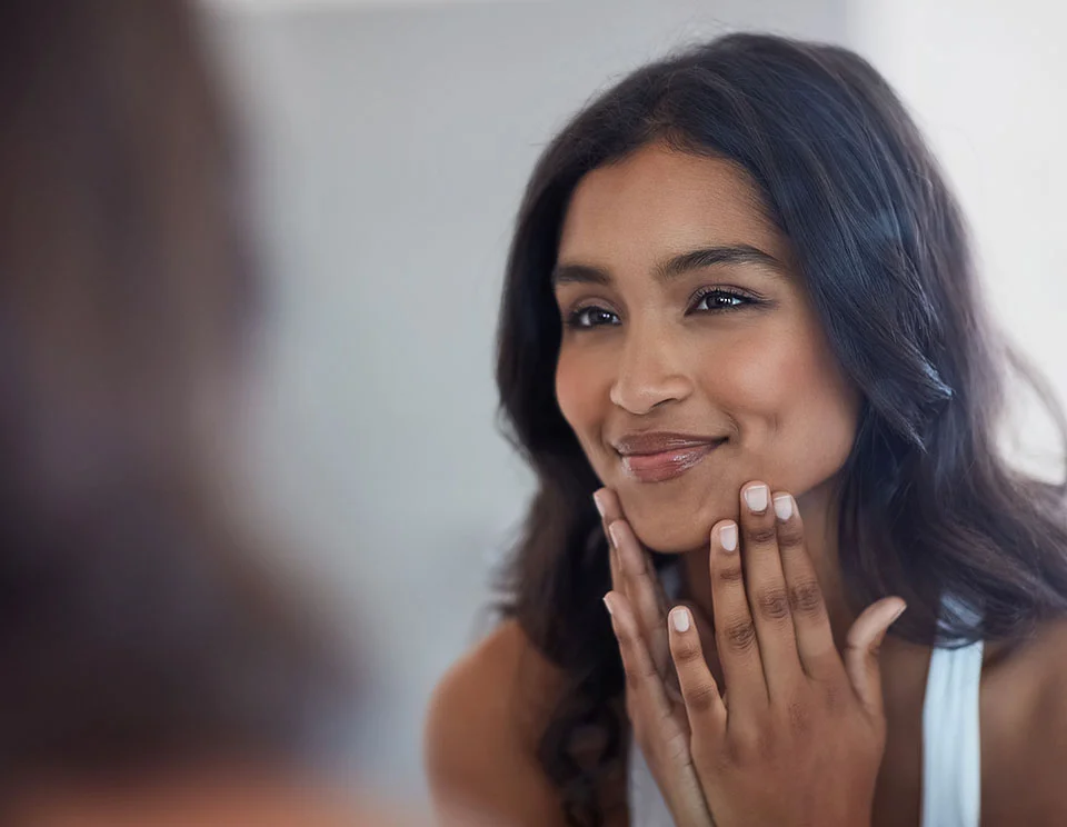 A young woman with long, wavy black hair, gently touching her face and admiring her reflection in the mirror. She has a soft, pleased expression on her face - Lip Lift in Portsmouth, NH