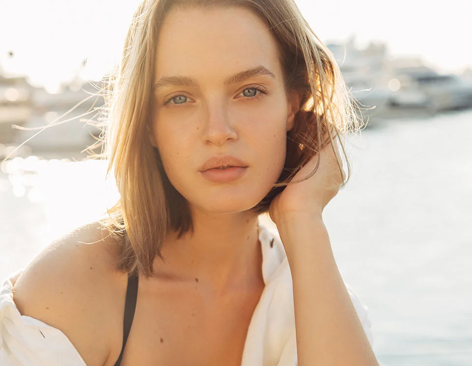 A close-up portrait of a woman with short, blonde hair, gently resting her hand on her face while gazing at the camera, with a soft, glowing background of yachts and water - Mole Removal in Portsmouth, NH