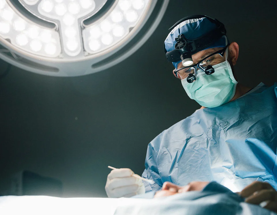 Surgeon in a blue surgical gown and mask performing a medical procedure under bright operating lights in a sterile operating room - Revision Rhinoplasty in Portsmouth, NH