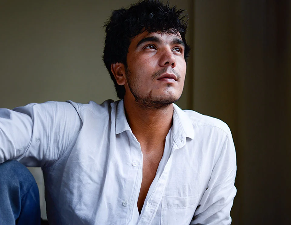 A young man with dark hair and a rugged look, dressed in a white button-down shirt, gazing thoughtfully upward - Top Surgery in Portsmouth, NH